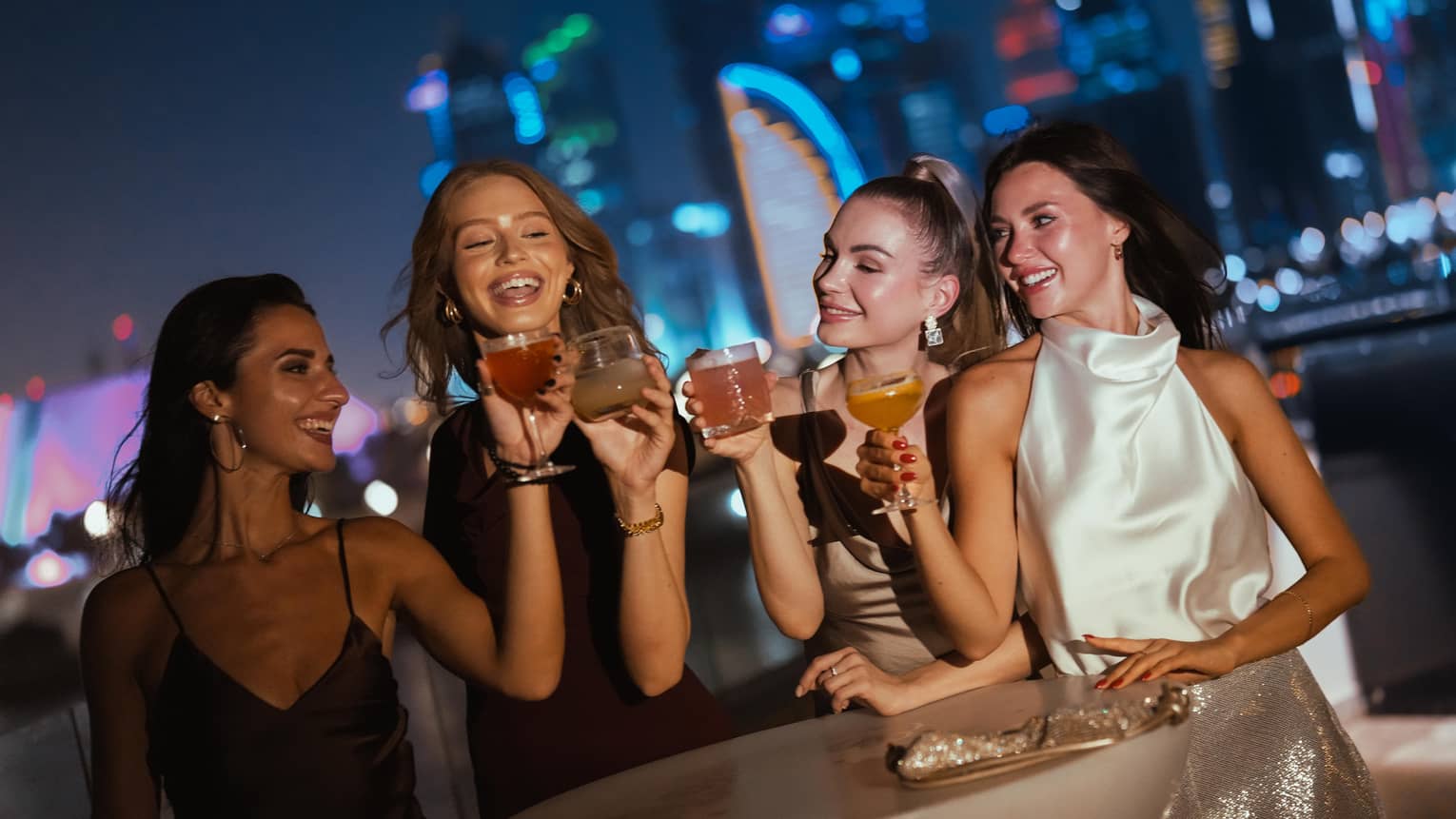 Four guests dressed in cocktail dresses stand at a rooftop dining table each holding a drink in one hand and smiling