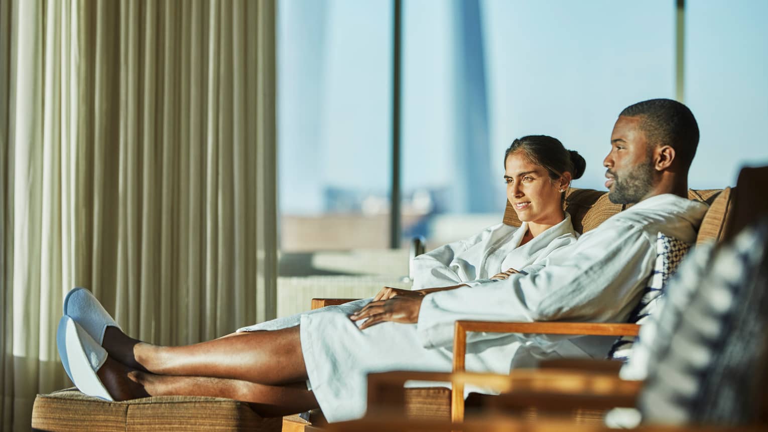 Couple wearing white bathrobes lounge near window, legs elevated on chair 