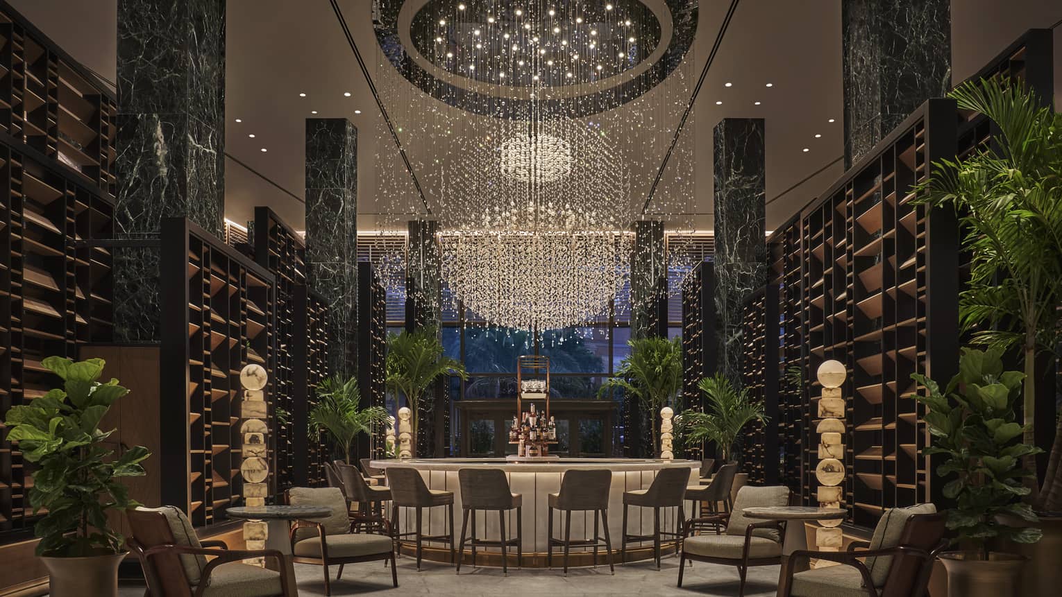 Chandelier Bar with six jade pillars flanked by wooden shelves and potted palms, glass-topped bar in the middle with six cocktail chairs, giant chandelier hanging overhead