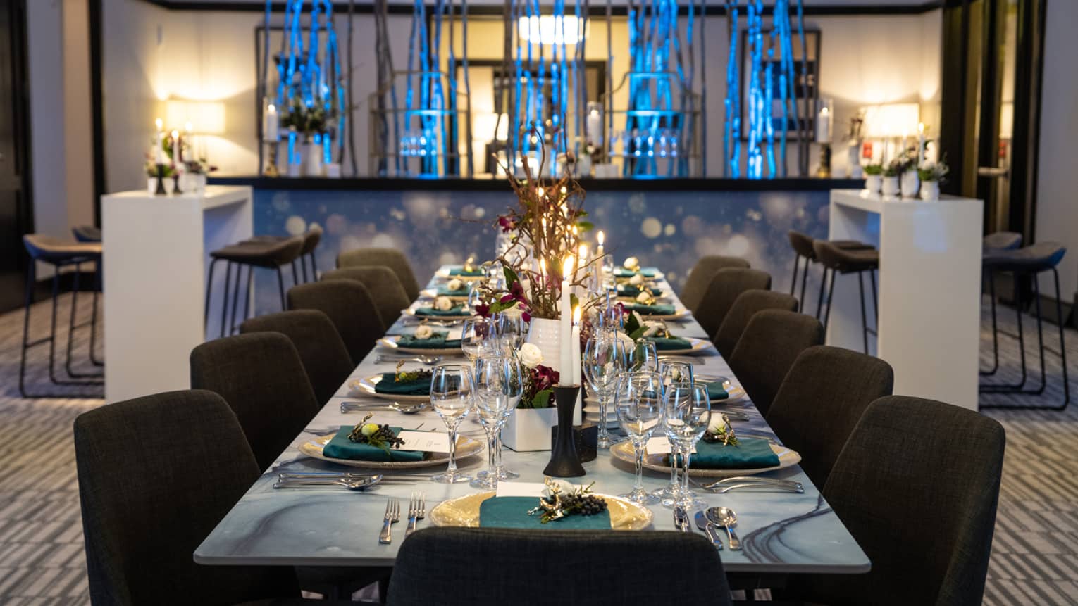 A long grey dining table with black chairs, teal napkins, and white plates; a bar is behind the table.