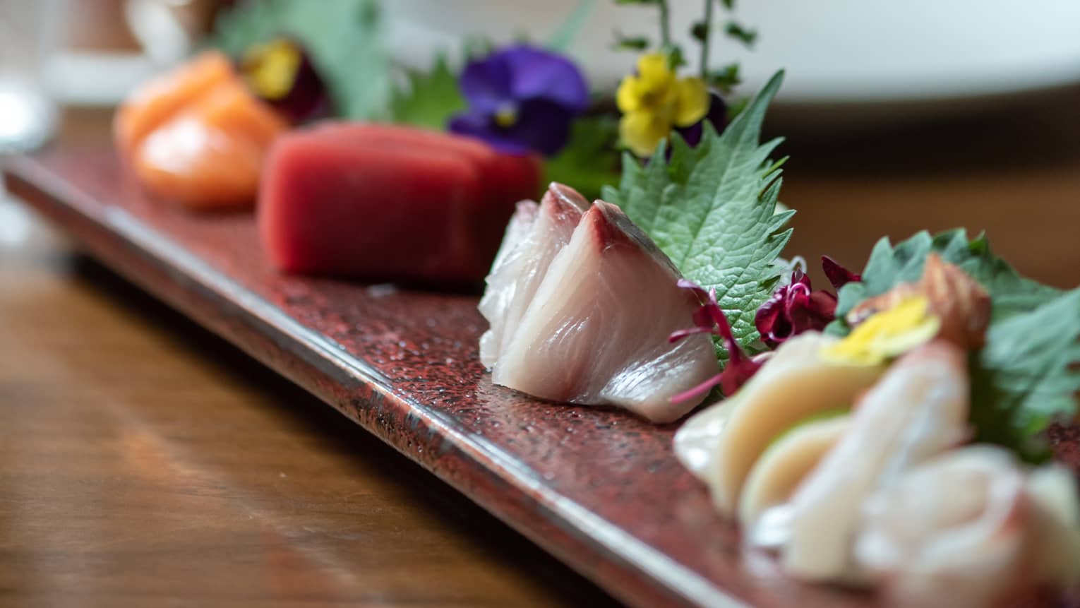 A long plate of multicoloured sashimi artfully adorned with delicate flowers; in the background, chopsticks poised overhead.
