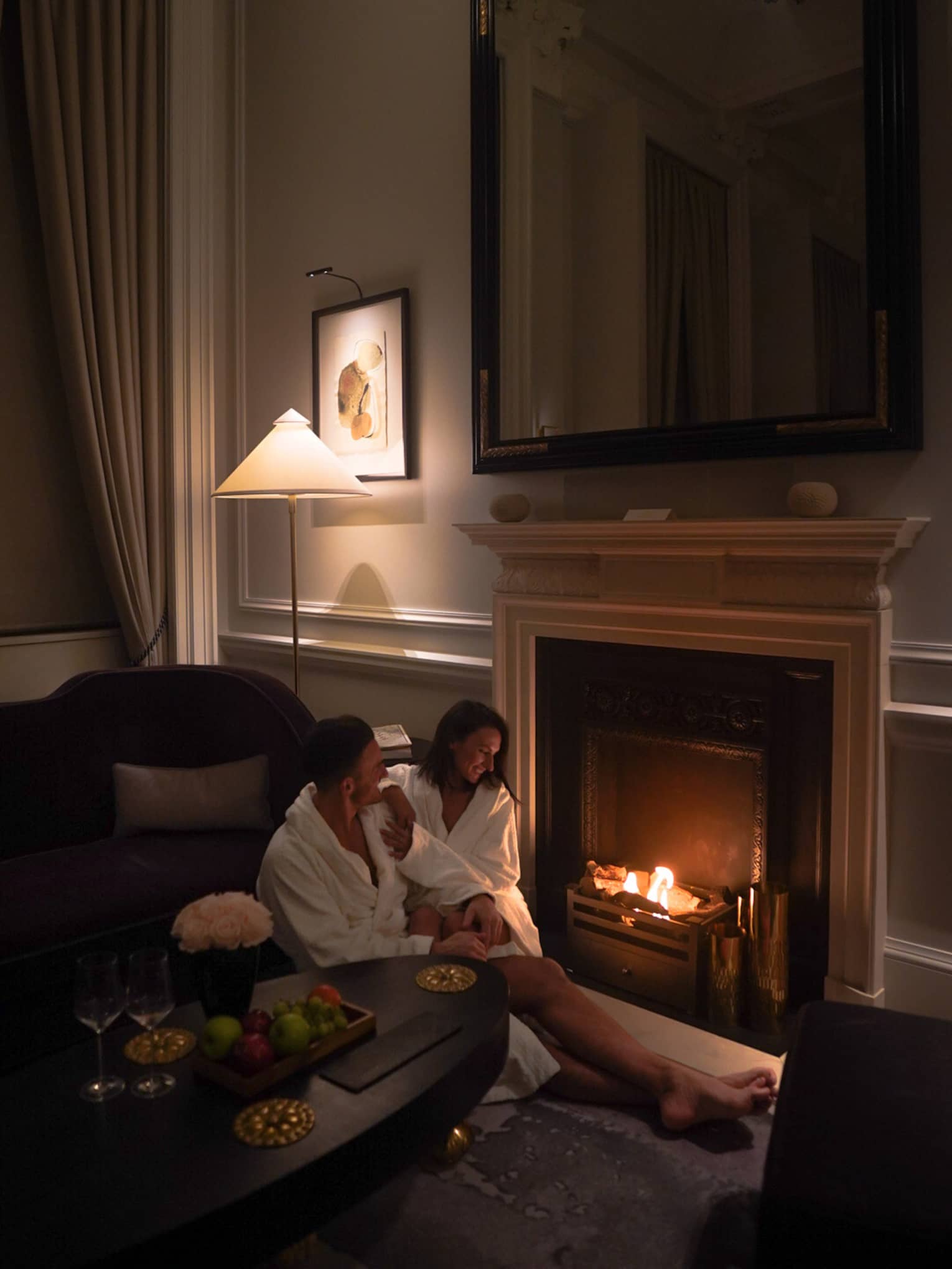 A couple dressed in white bath robes lounges next to a fireplace in a dimly lit room