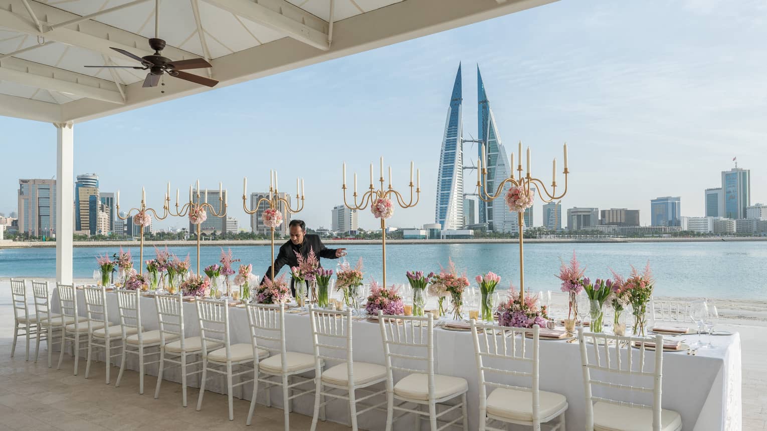 Hotel staff sets long wedding dining table by waterfront, city skyline in background