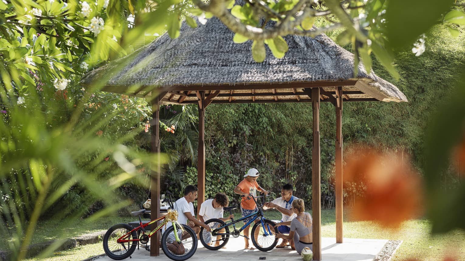 View through foliage of four adults and a child working on a bicycle under a thatched hut surrounded by lush greenery.