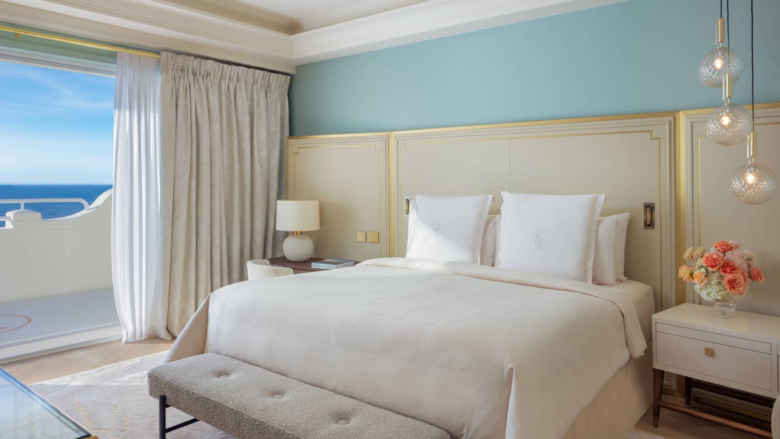 Elegant luxury hotel suite bedroom with king bed, ivory bedding and headboard with gold accents and turquoise wall, with walk-out sea-view balcony