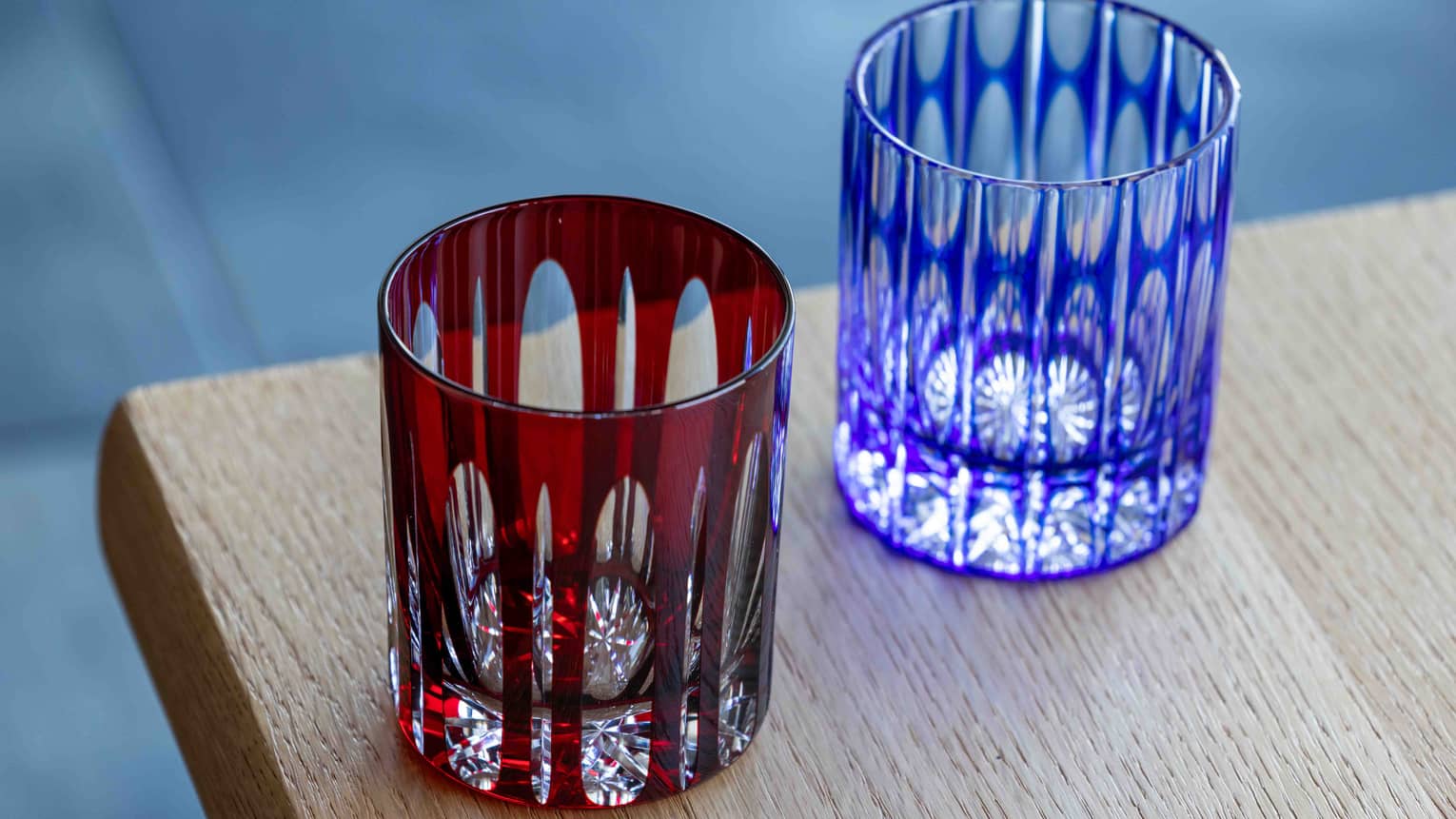 Close-up of two cut-glass tumblers with intricate geometric patterns overlaid by coloured glass, one dark red, one blue.
