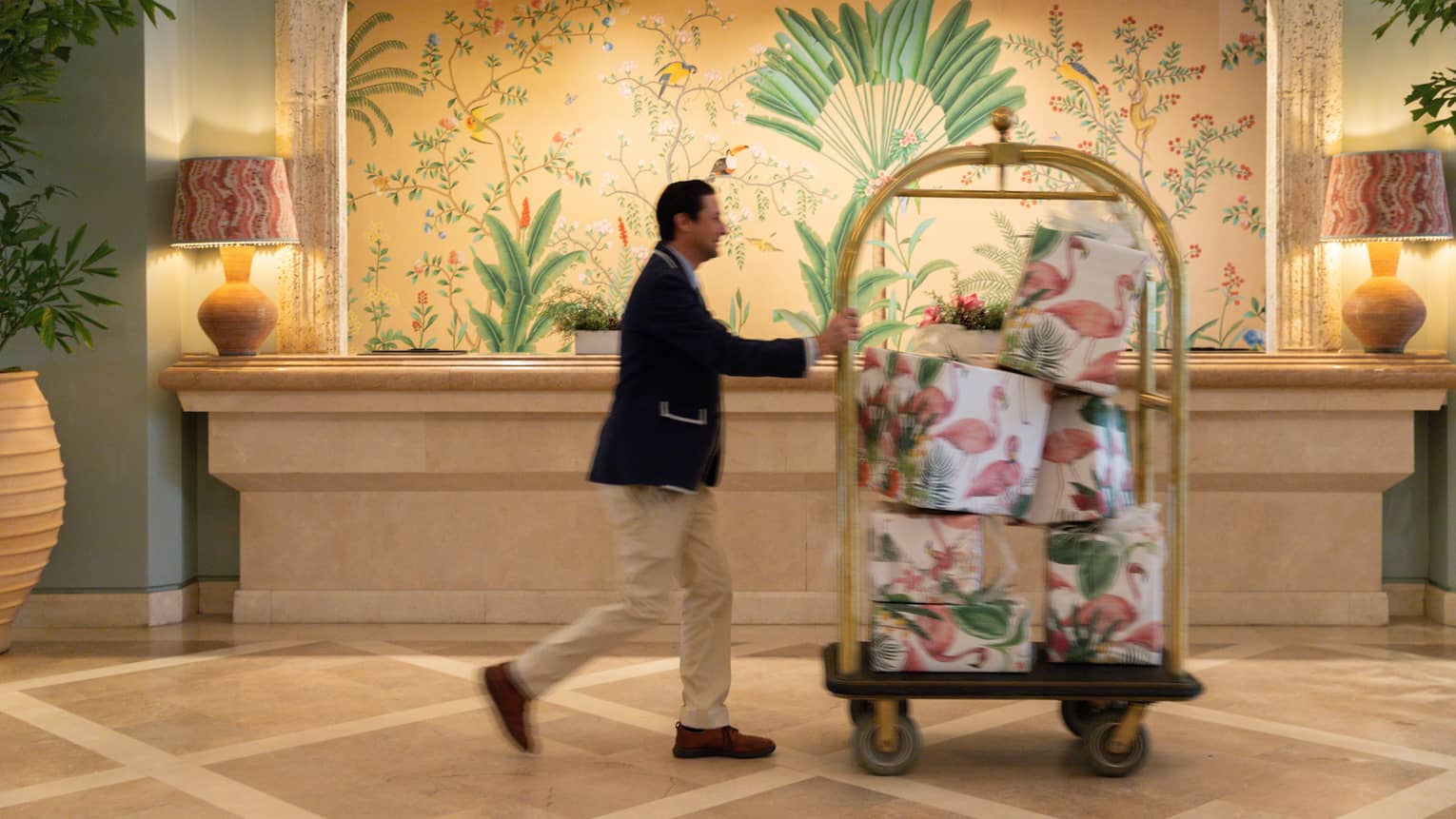 Bellhop wearing navy blazer and khaki pants pushes a luggage cart filled with presents through a hotel lobby