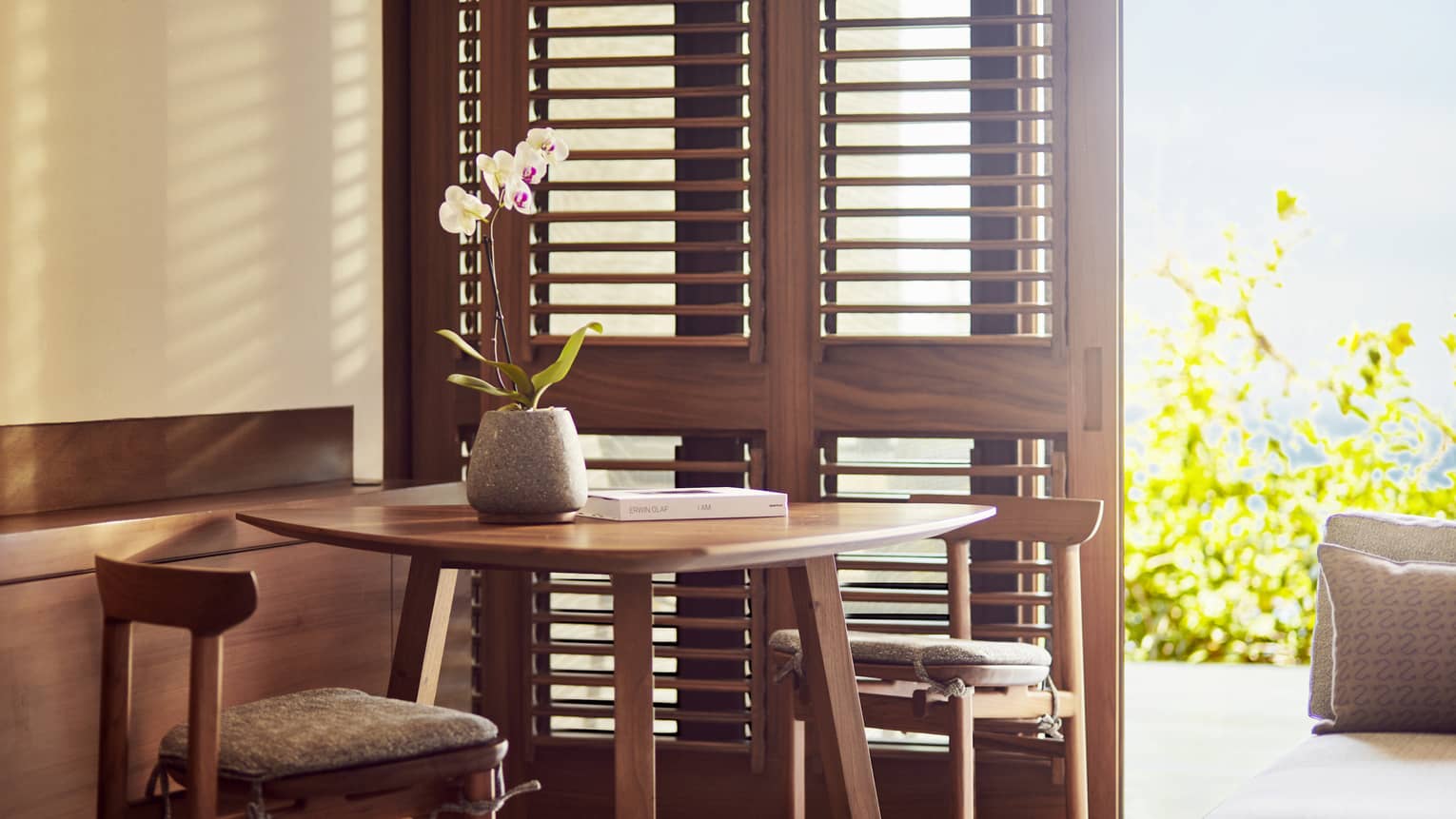 Resort bedroom with wooden table and two chairs next to shutter doors opening to terrace