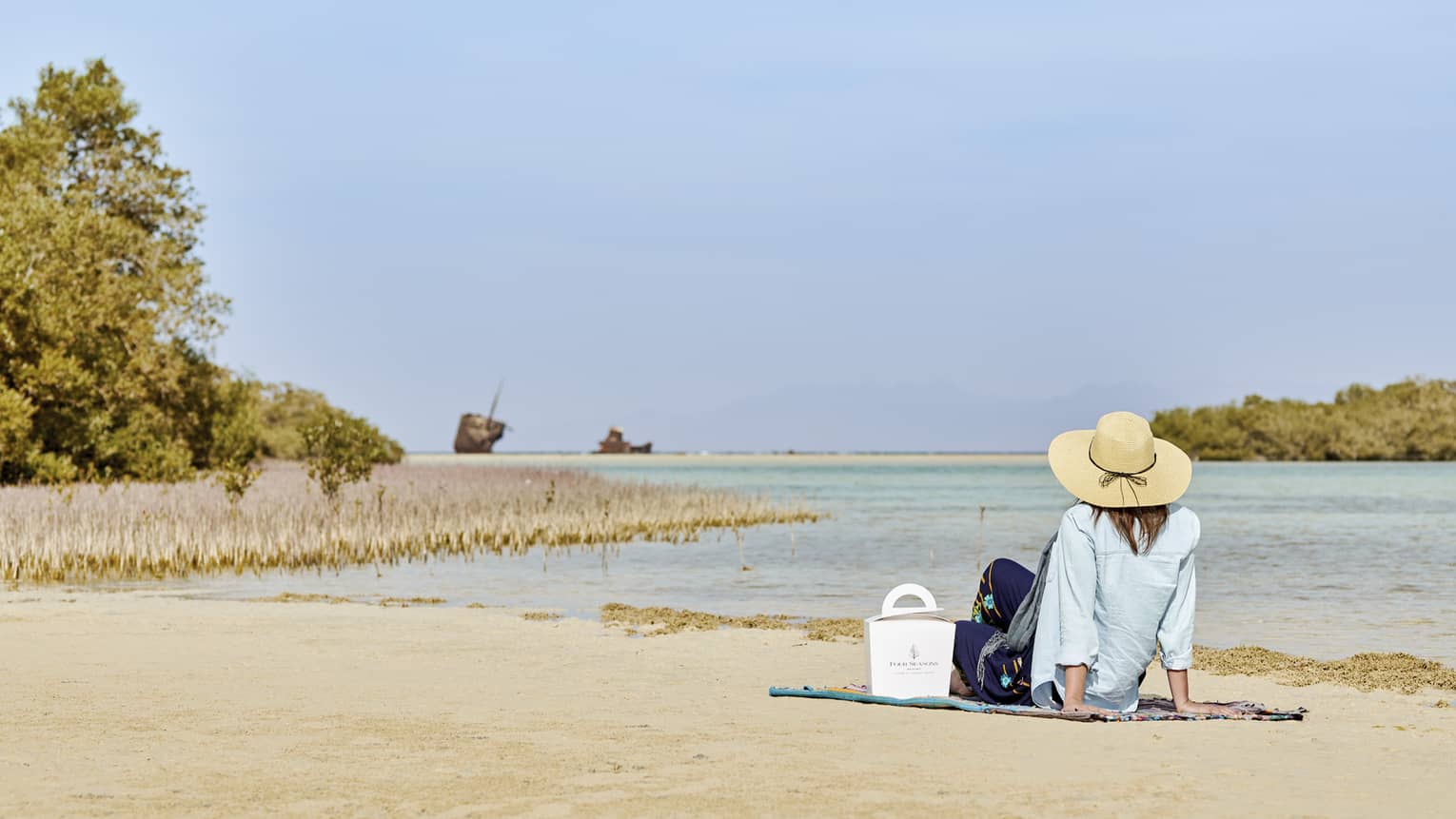 A guest in a sunhat, a Four Seasons food container beside them, sits on a beach by the water's edge, gazing at the horizon.