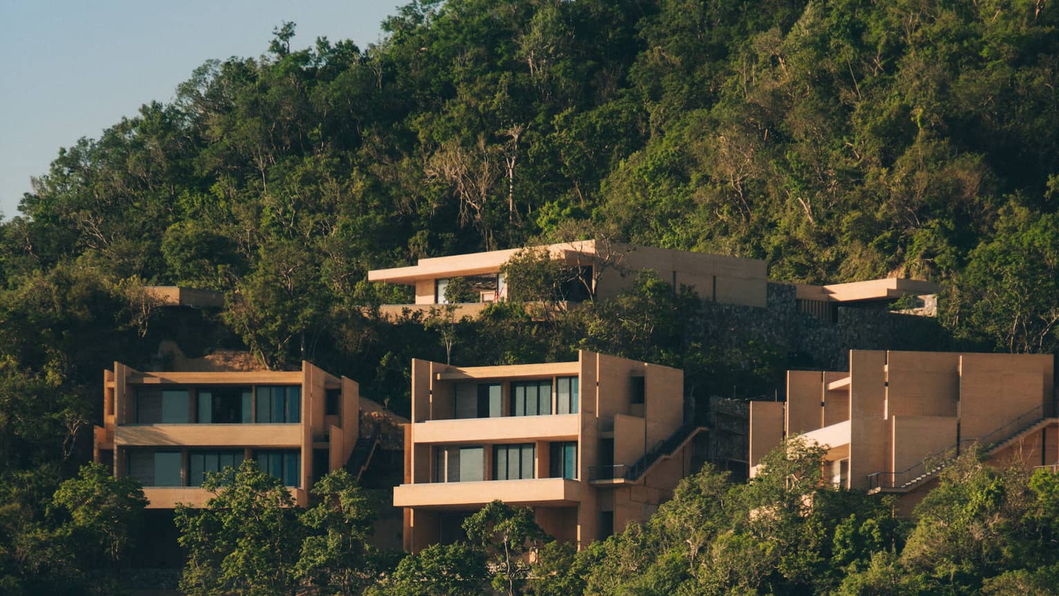 Modern cliffside villas surrounded by lush greenery, overlooking a secluded beach and ocean. The homes feature clean, angular designs, blending into the natural landscape.