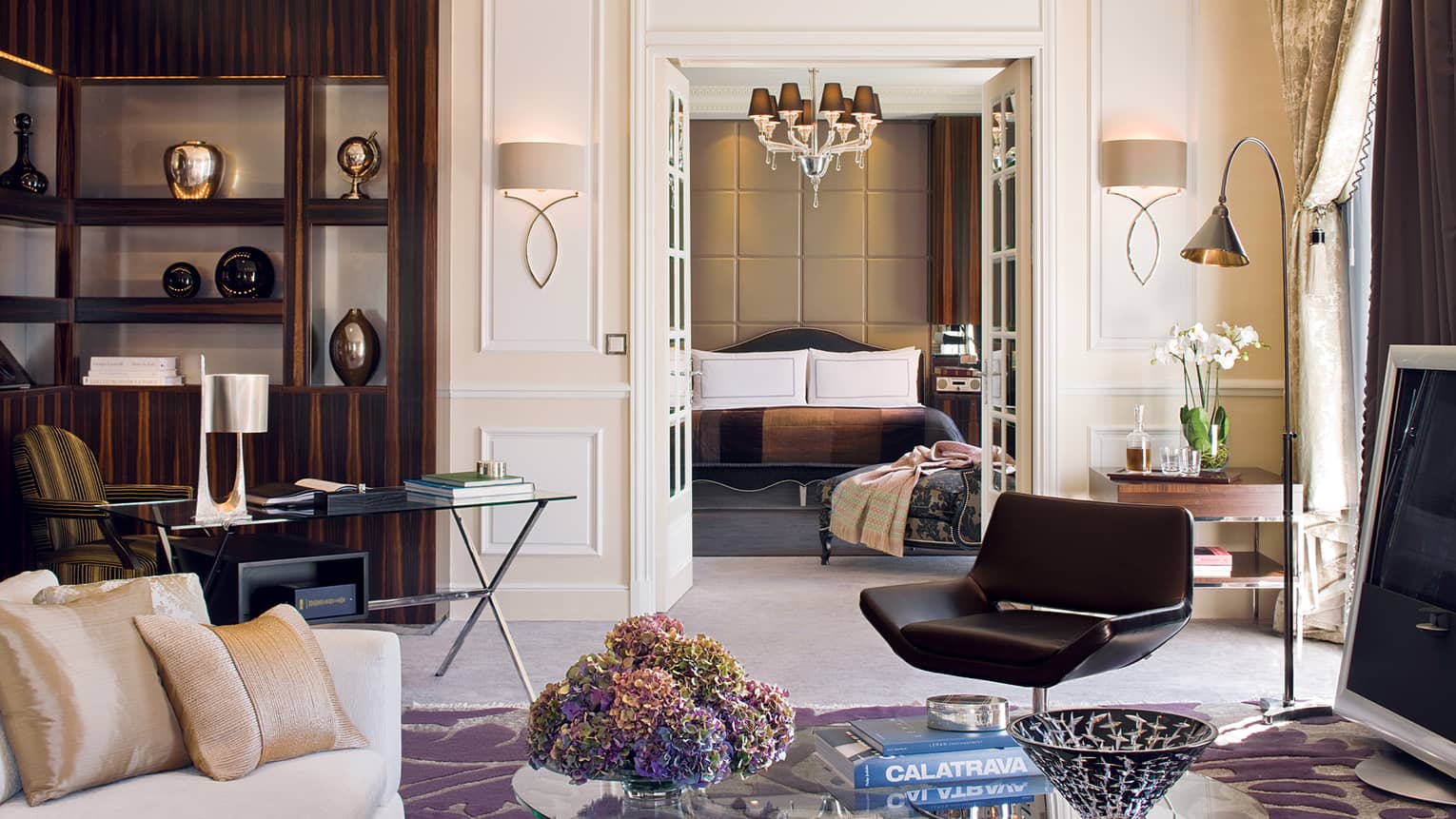 Presidential suite living area with sleek dark furniture, a glass coffee table, beige sofa, desk and decorative shelves, leading into a luxury bedroom