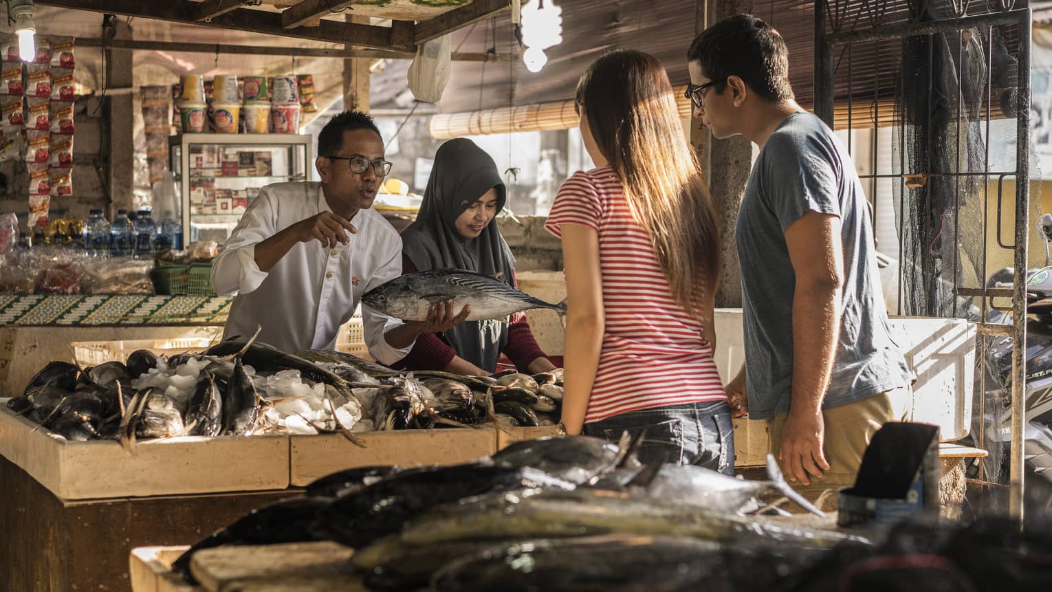 A merchant presents a fish to two tourists before a table of fish in a bed of ice inside a covered market. 