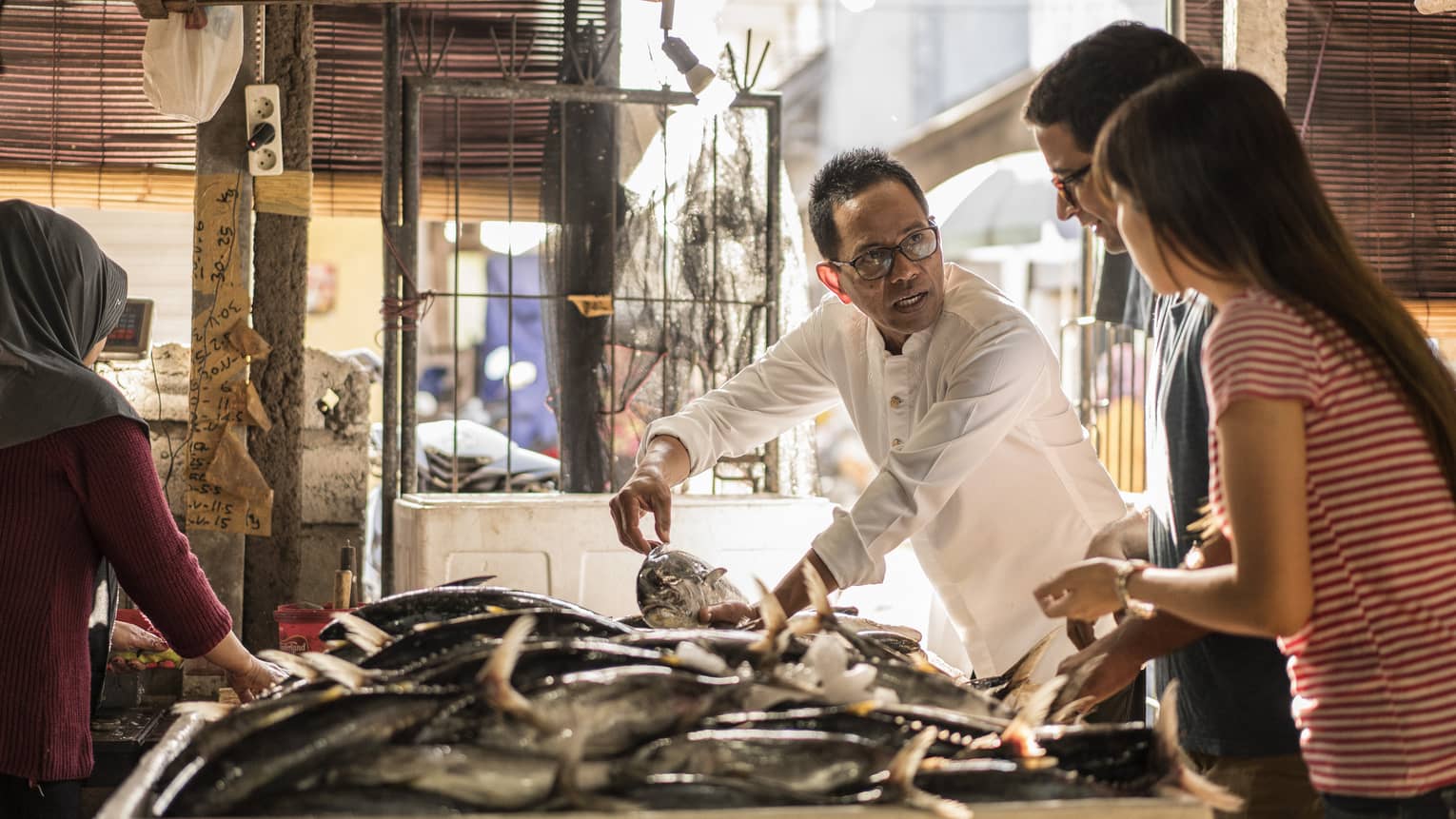 A merchant gently holds a fish with two hands while talking to two guests as they all stand by an indoor market fish table.
