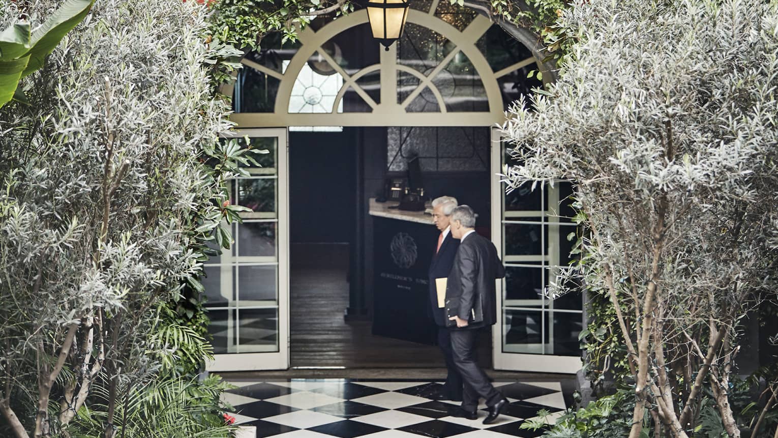 Two man wearing business suits walk by arched door, window in hotel courtyard