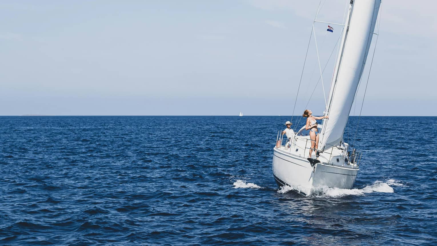 Front view of two guests aboard a sailboat as it leans into an expanse of dark blue waters under an azure cloudless sky.