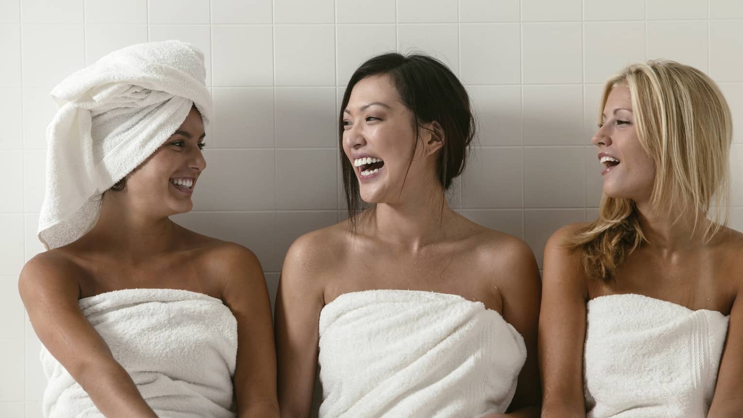 Three people in a spa setting, wrapped in white towels, smiling and laughing. One has a towel around their hair. The background features a tiled wall.