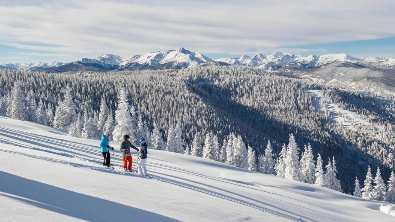 Three guests on skis pause on the ski hill, snow capped trees and mountains in distance