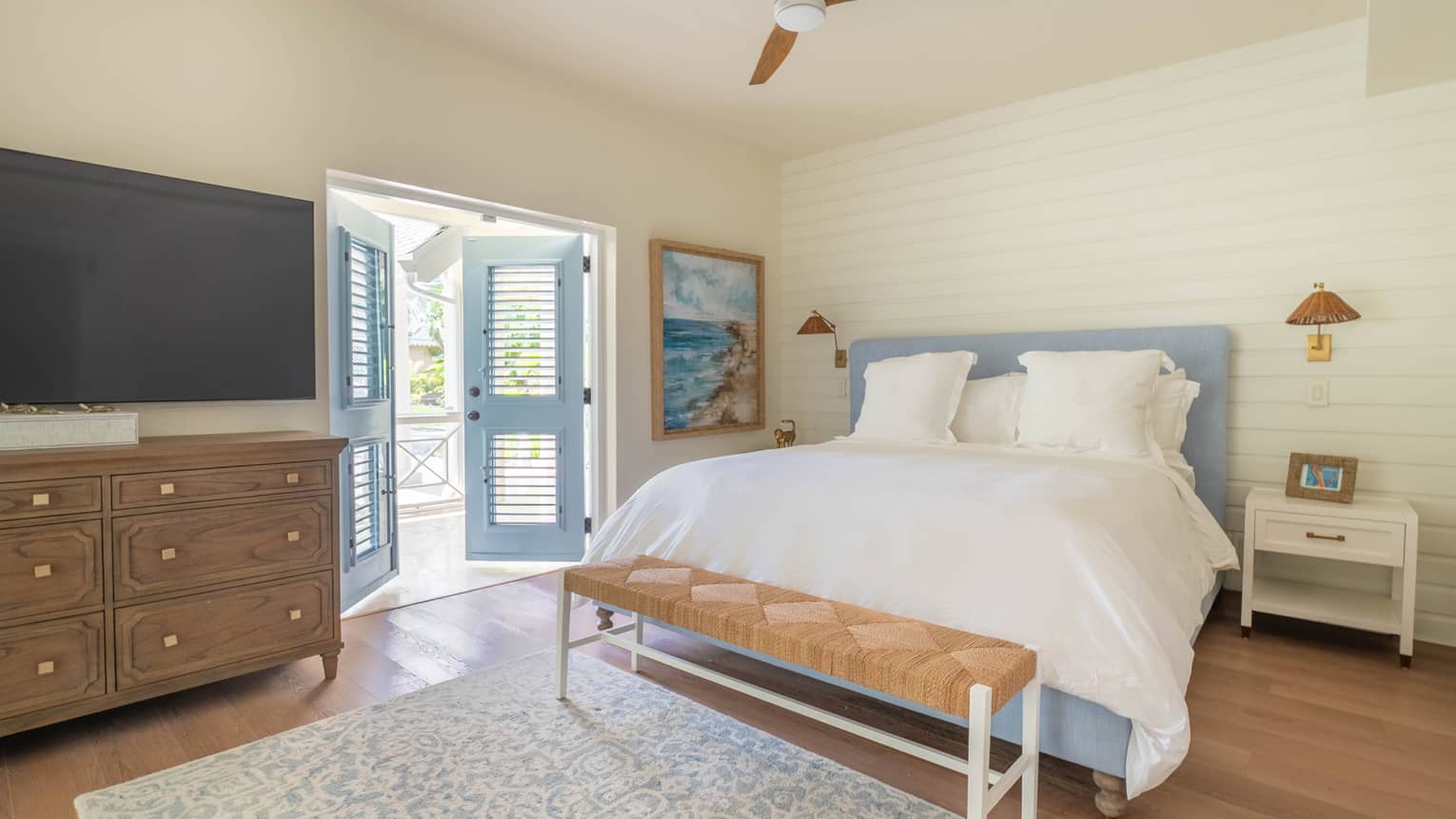 Bedroom with king bed, wood floors, blue and white rug, ceiling fan, dresser, TV and blue shuttered doors leading to outside