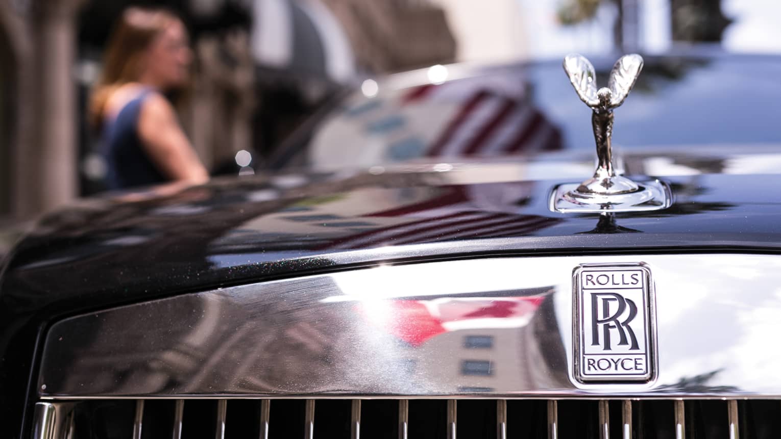 Close-up view of a winged silver ornament on the hood of black Rolls Royce, double-R logo on reflective chrome above grille.