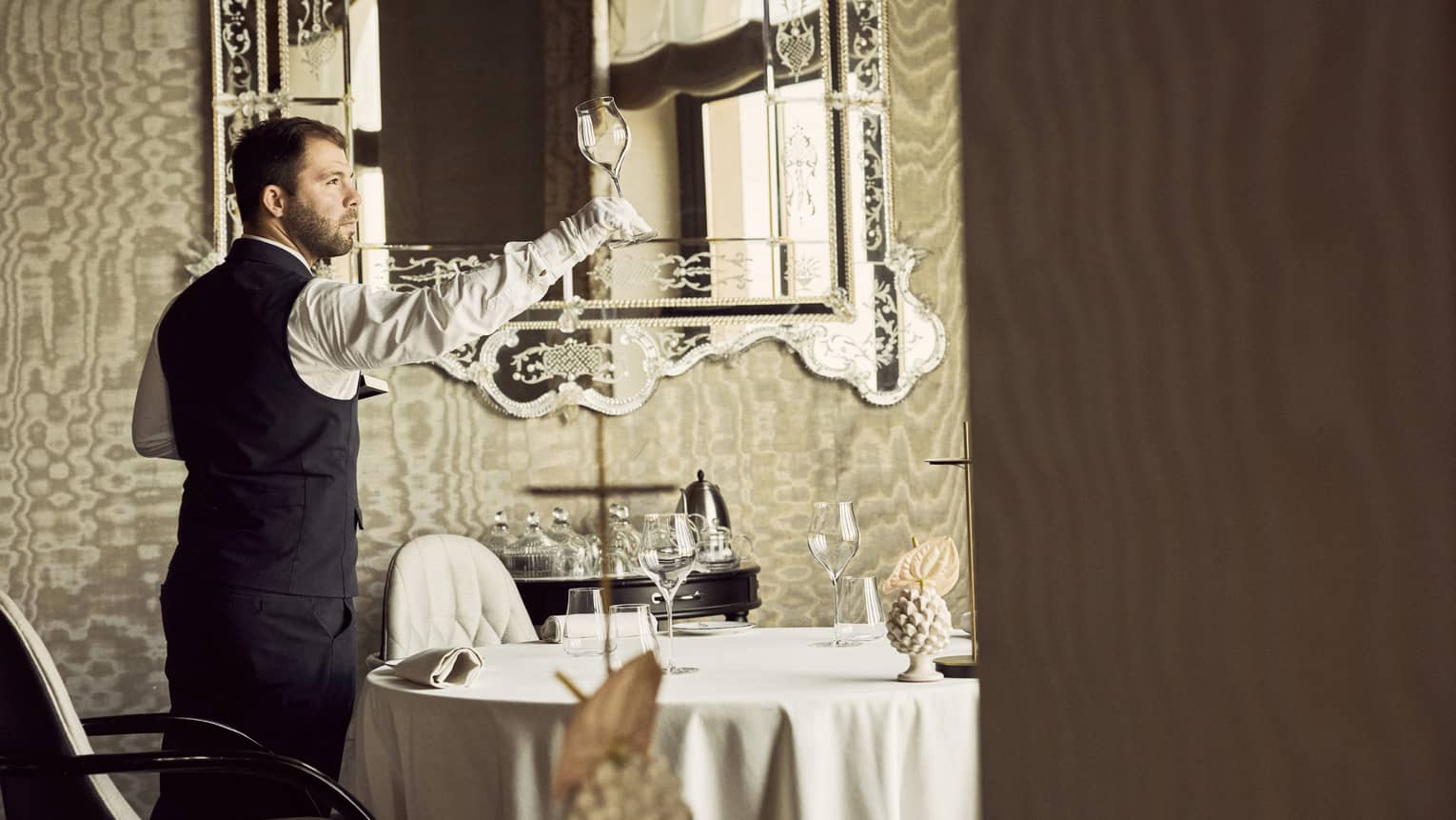 Server wearing black pants, black vest, white long-sleeve button down shirt and white gloves inspects an empty wine glass in a dining room with a large ornate mirror behind them