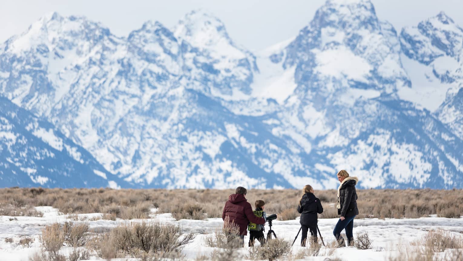 A family in winter apparel uses two telescopes to view a snowy mountain peaks