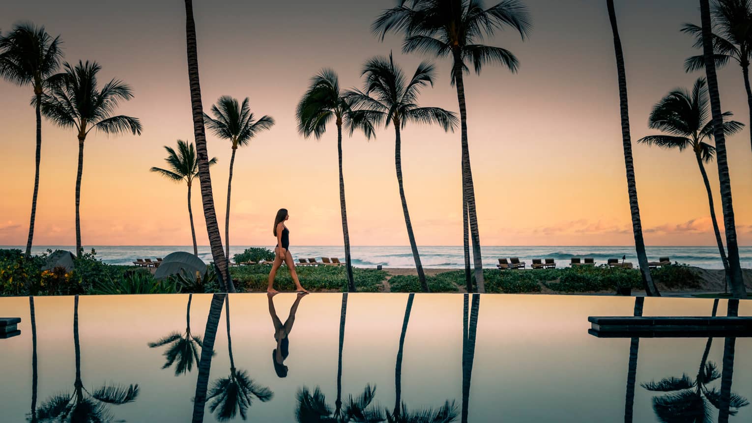 Woman in swimsuit walks by Palm Grove Pool at sunset, palm trees reflected on surface