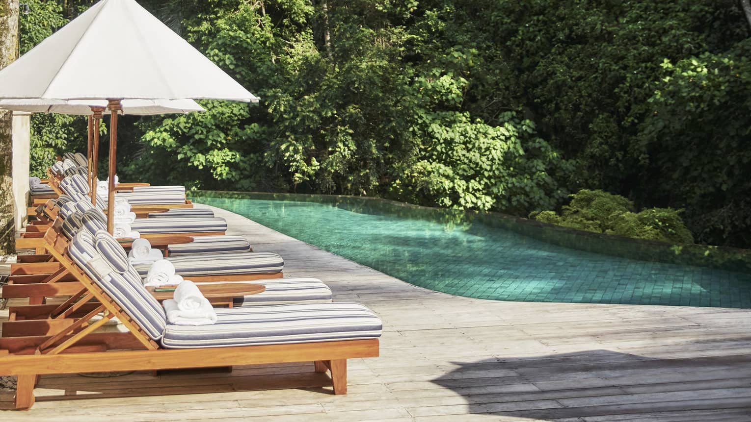 Eight pool lounge chairs with striped cushions, white umbrella lined up near blue swimming pool on sunny day 