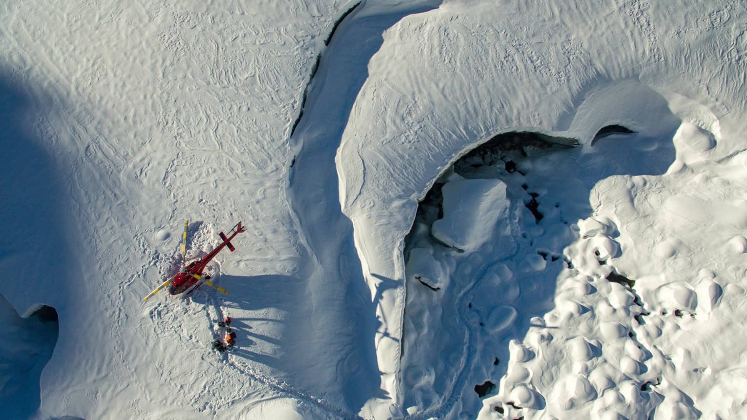 Aerial view of a red helicopter that's landed on a snow-covered mountain with three people walking near it