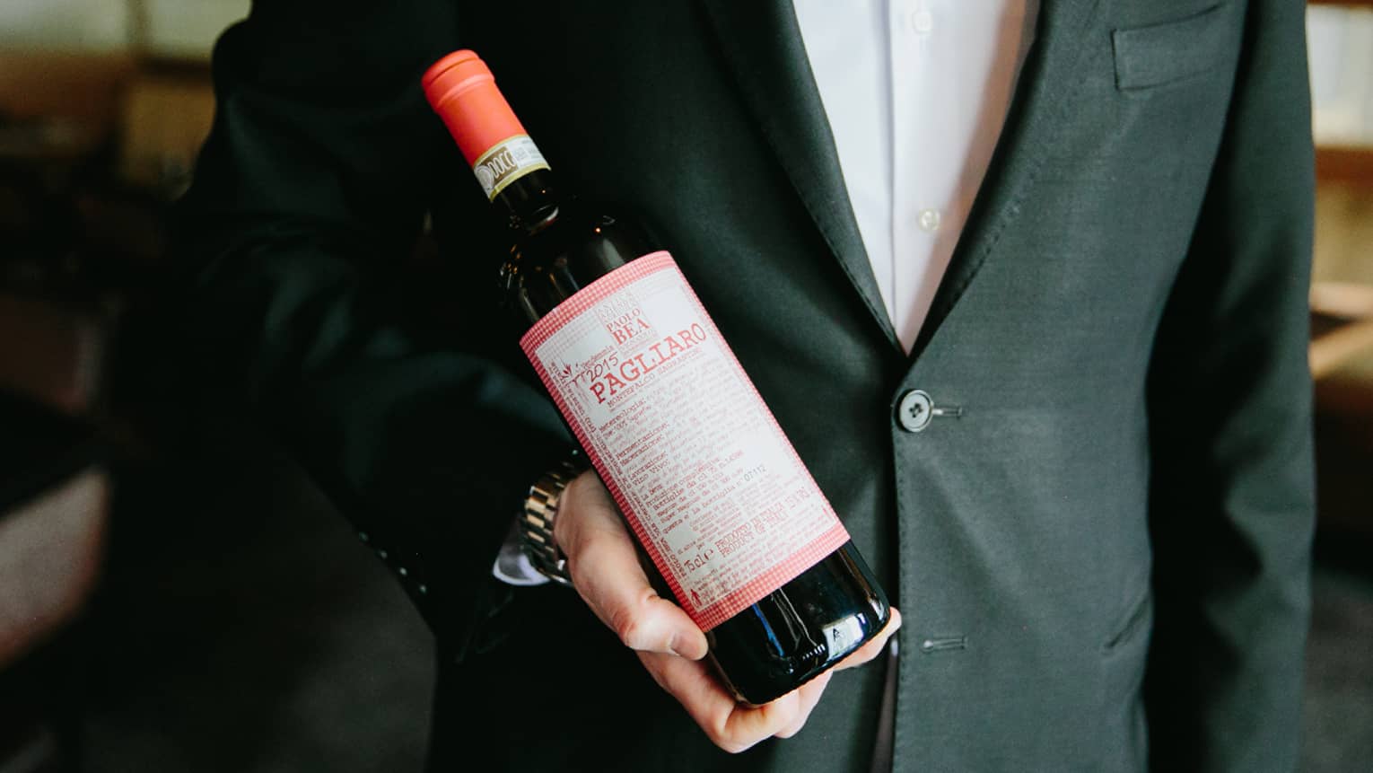 A man dressed in a suit holds a bottle of wine with a red label