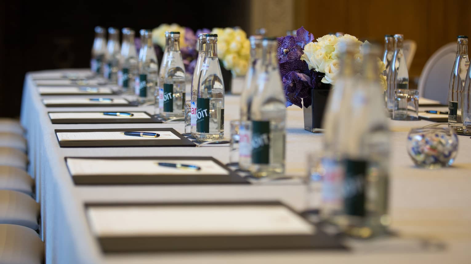 Row of meeting agendas, glass bottles, small vases with flowers on boardroom table