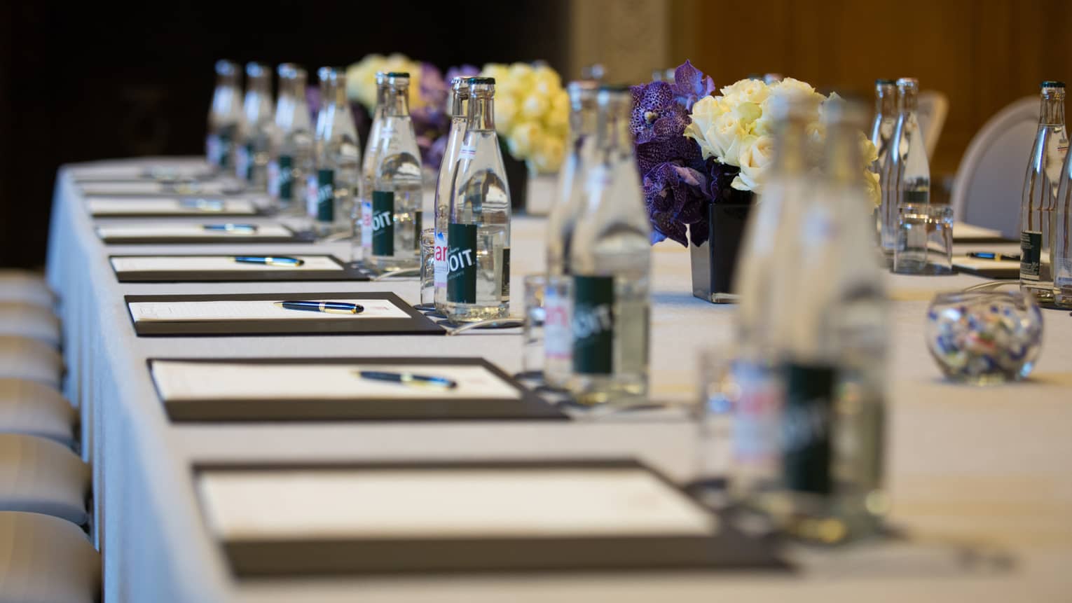 Row of meeting agendas, glass bottles, small vases with flowers on boardroom table