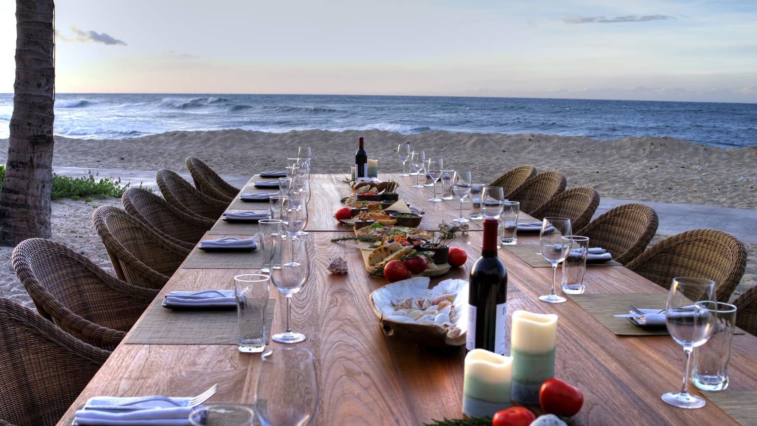 View down long wood private dining tale with wine, candles and shells, wicker chairs on beach