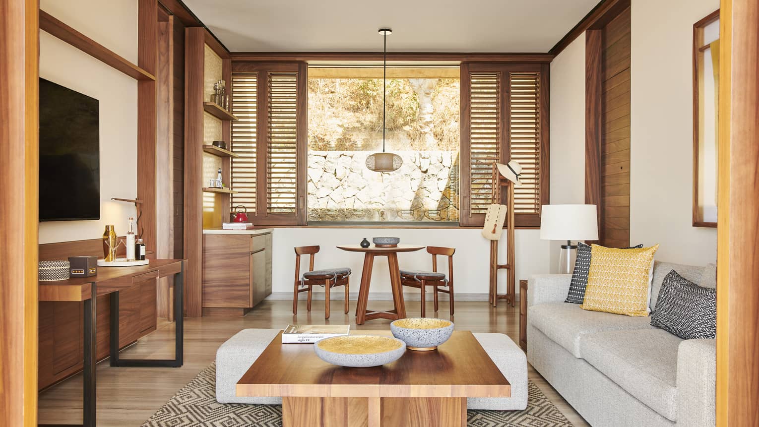 Stylish living area with natural wood furniture, a cozy grey sofa and a small dining table. The room features large windows with shutters, offering a view of a stone wall outside.