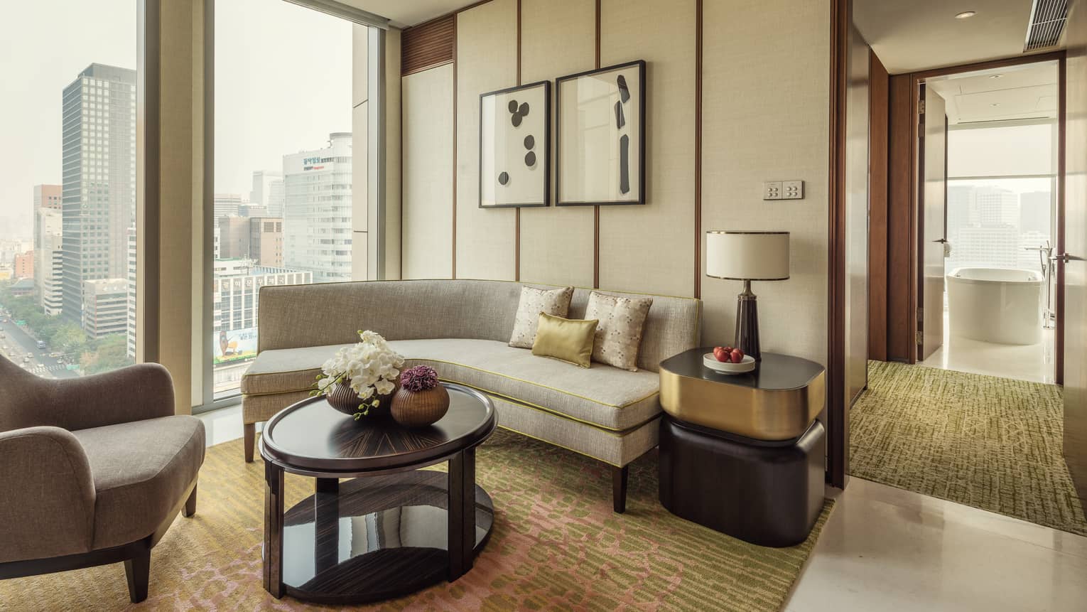 City-View Executive Suite curved sofa under window, armchair, table with lamp by hallway to bathroom