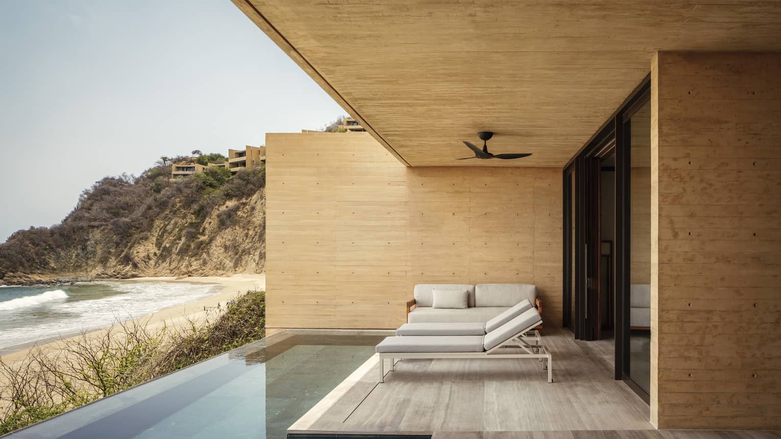 Luxury outdoor patio with minimalist furniture and an infinity pool, overlooking a secluded sandy beach and rugged cliffs. The design features natural wood and modern accents.