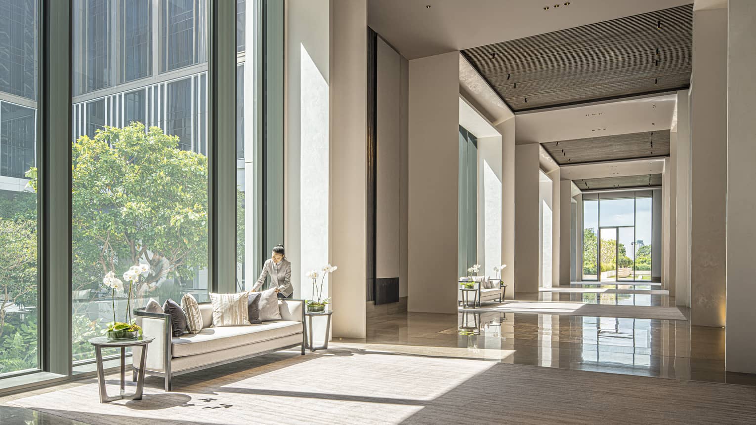 Staff member fluffs the couch pillows in the Ballroom Foyer with cream-coloured carpeting and furniture, floor-to-ceiling windows