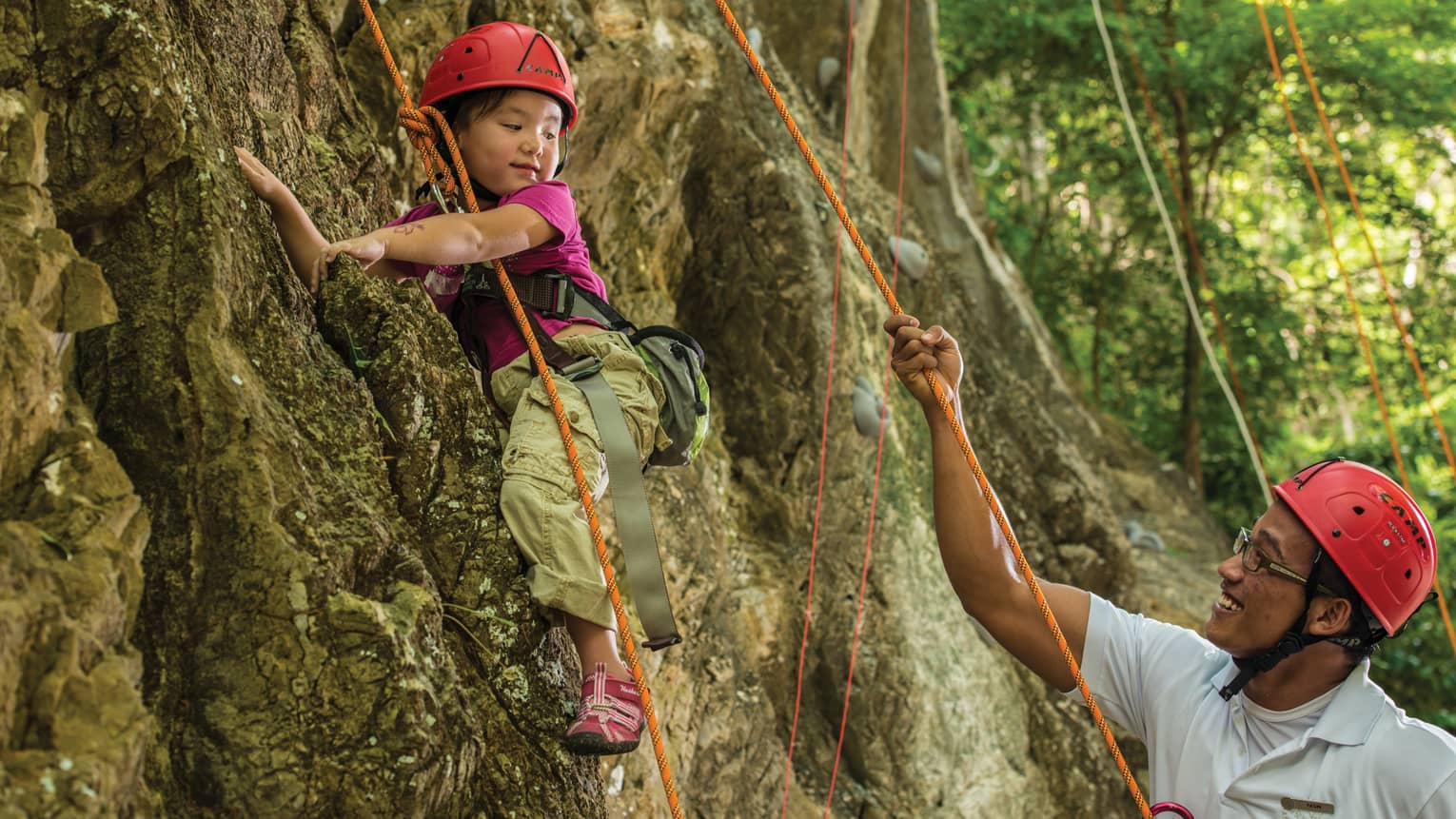 Man holds rope, helps young girl wearing helmet climb rock face