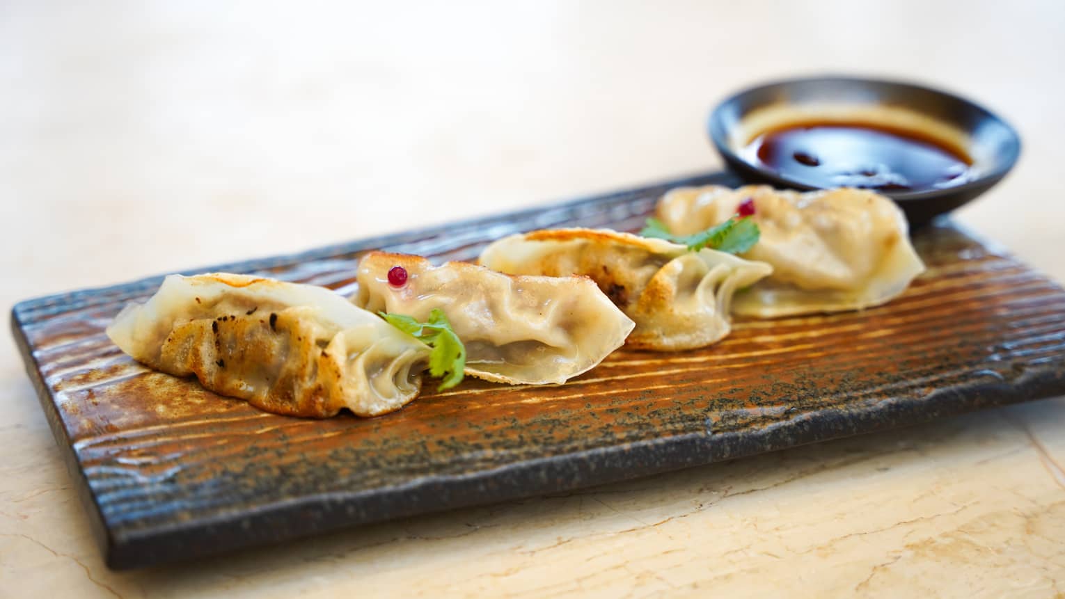 Row of four pan-fried dumplings on ceramic serving slab, small bowl of dipping sauce in the background