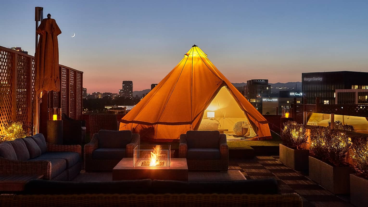 Close-up of illuminated white tent with bed, lamp by wicker chairs, modern outdoor fireplace