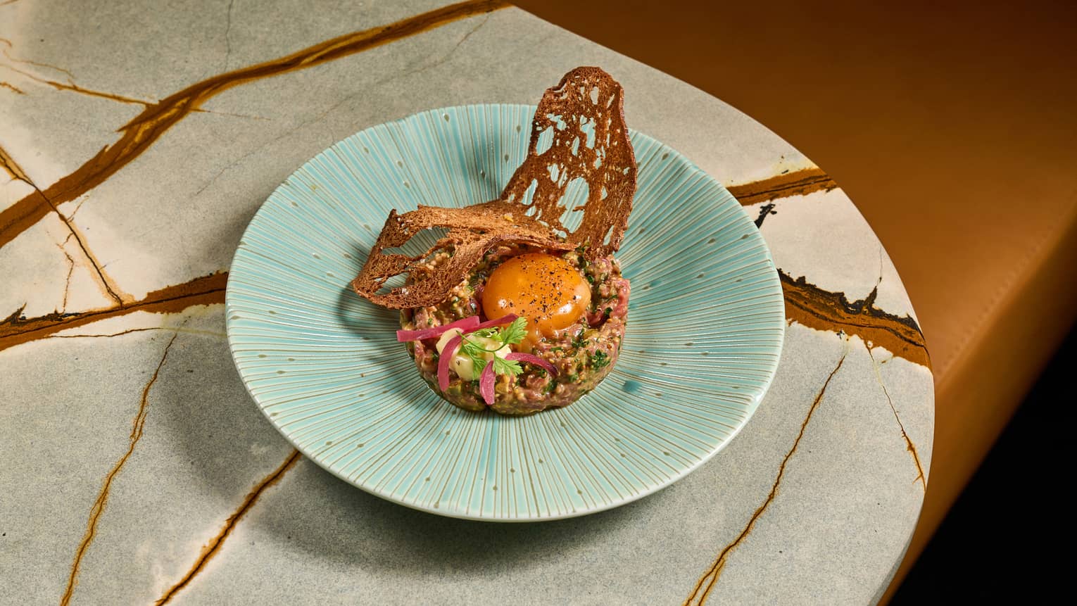  Cester beef tartare with dijion mustard ancient, cornichons, parsley, cured egg yolk and pickled onions served on a light-blue plate