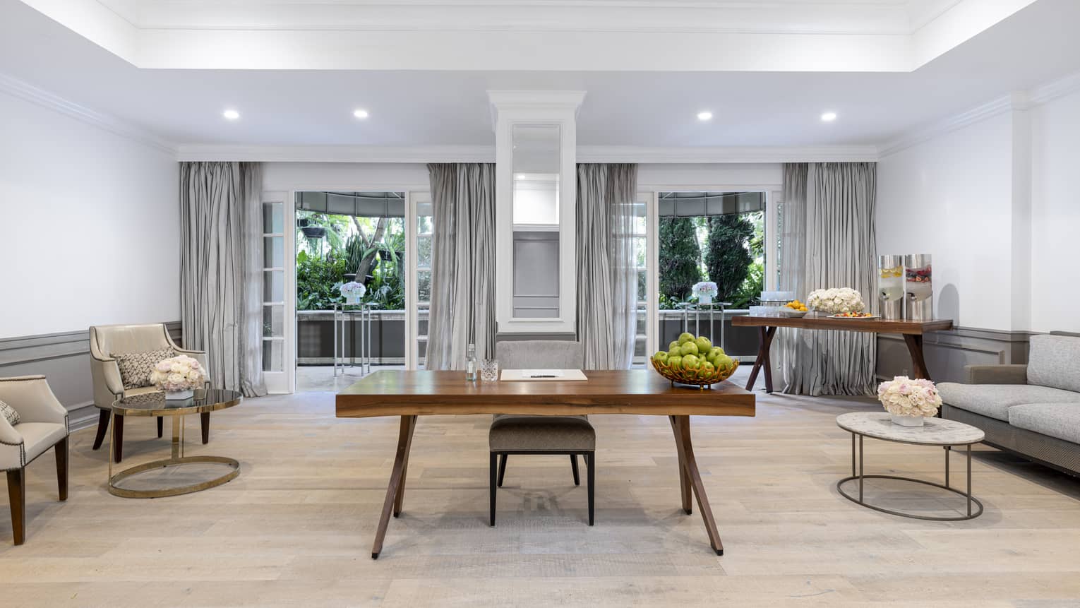 A seating room with a desk, two chairs, a sofa, a table with refreshments and large windows.