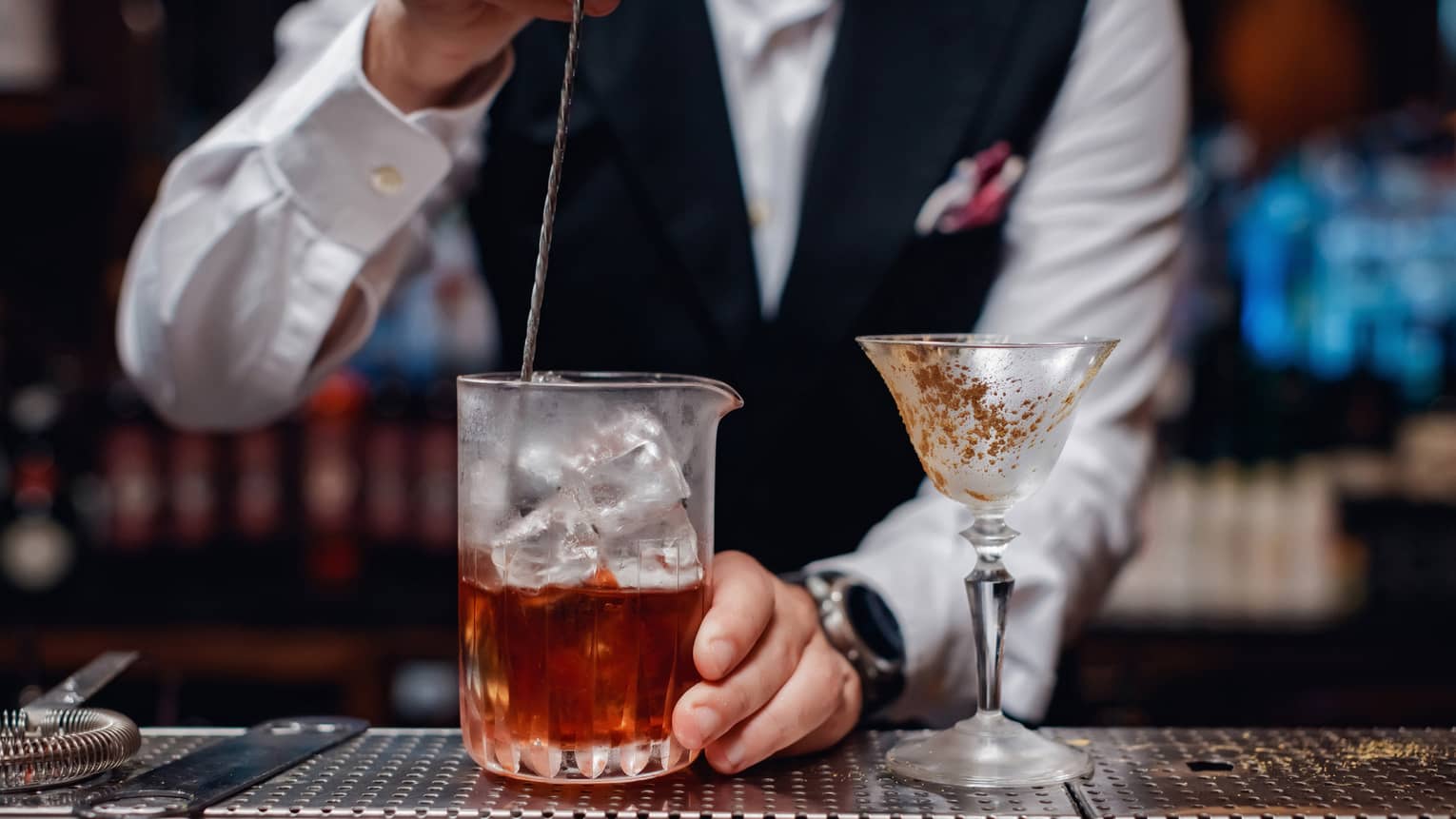 Close-up of bartender stirring liquor over ice in glass pitcher next to cocktail glass on bar 