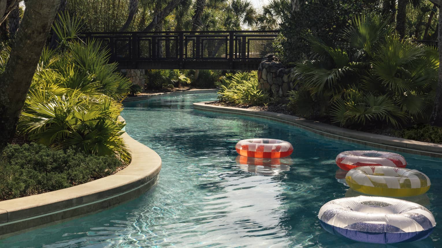 Colourful inflatable pool tubes float on a winding pool bordered by lush trees. A walking bridge over the pool is in the background.