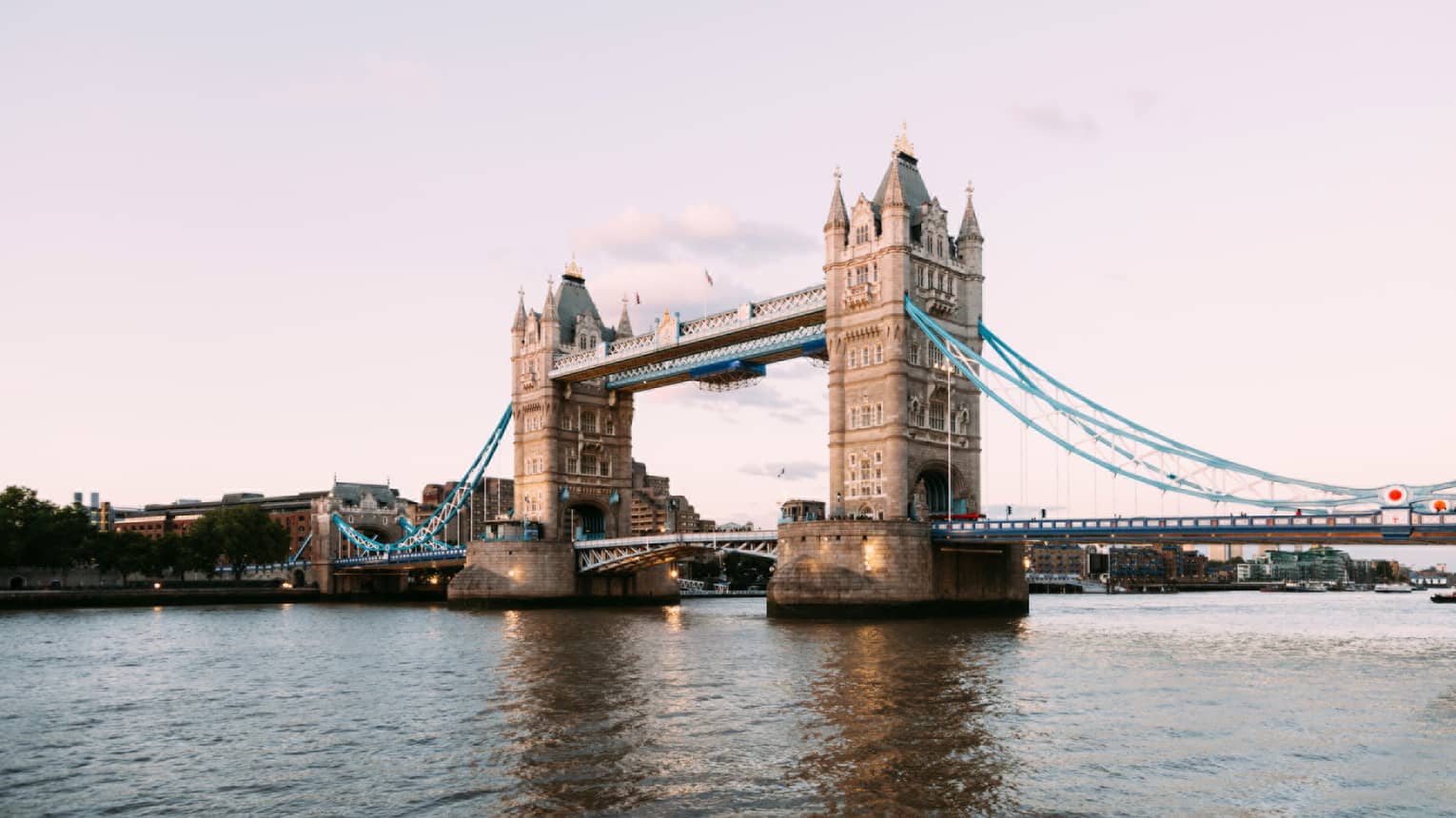 The iconic Tower Bridge of London spanning the dark Thames River in the foreground against a pale, dusky sky.