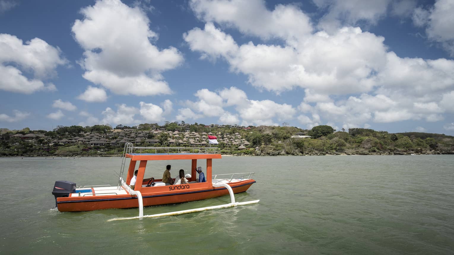 An outrigger canoe glides along calm, green water as four passengers look toward a distant shore dotted with trees and huts.