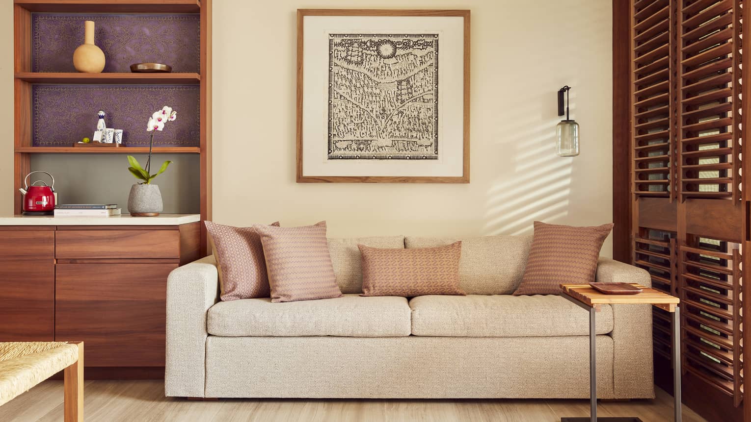A cozy living area with a beige sofa adorned with patterned pillows. A wooden shelf with decorative items, a framed artwork and natural light streaming through wooden shutters complete the space.