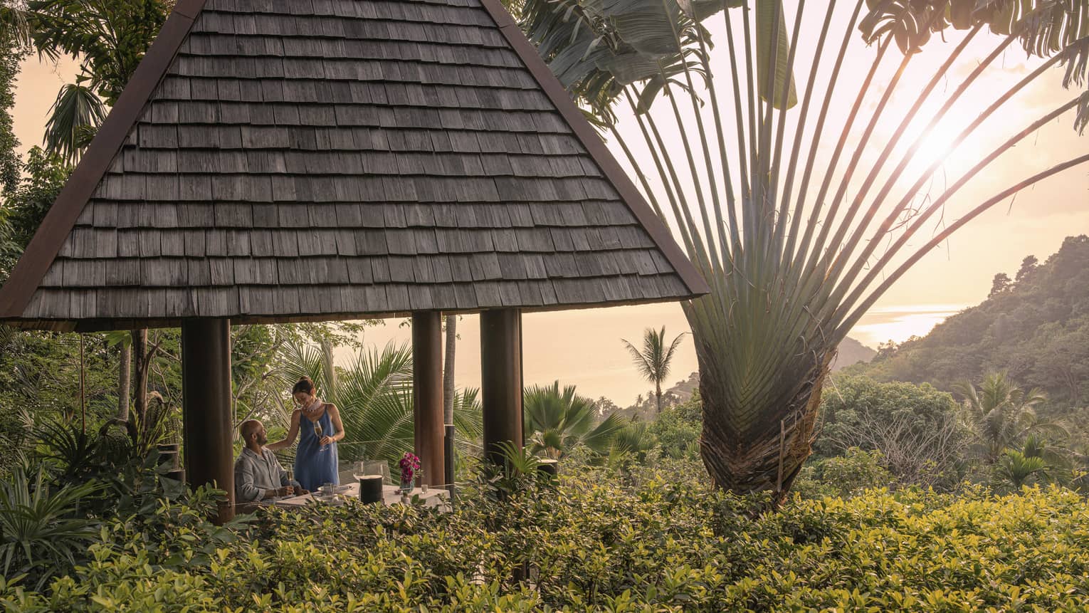 A couple with champagne in a tall hilltop pavilion with a dark, straw roof, set amidst lush forest and a giant fan palm tree.