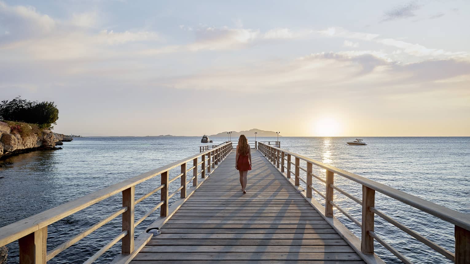 A woman walking along a long wooden walkway that extends into the ocean, the sun is setting in the background.