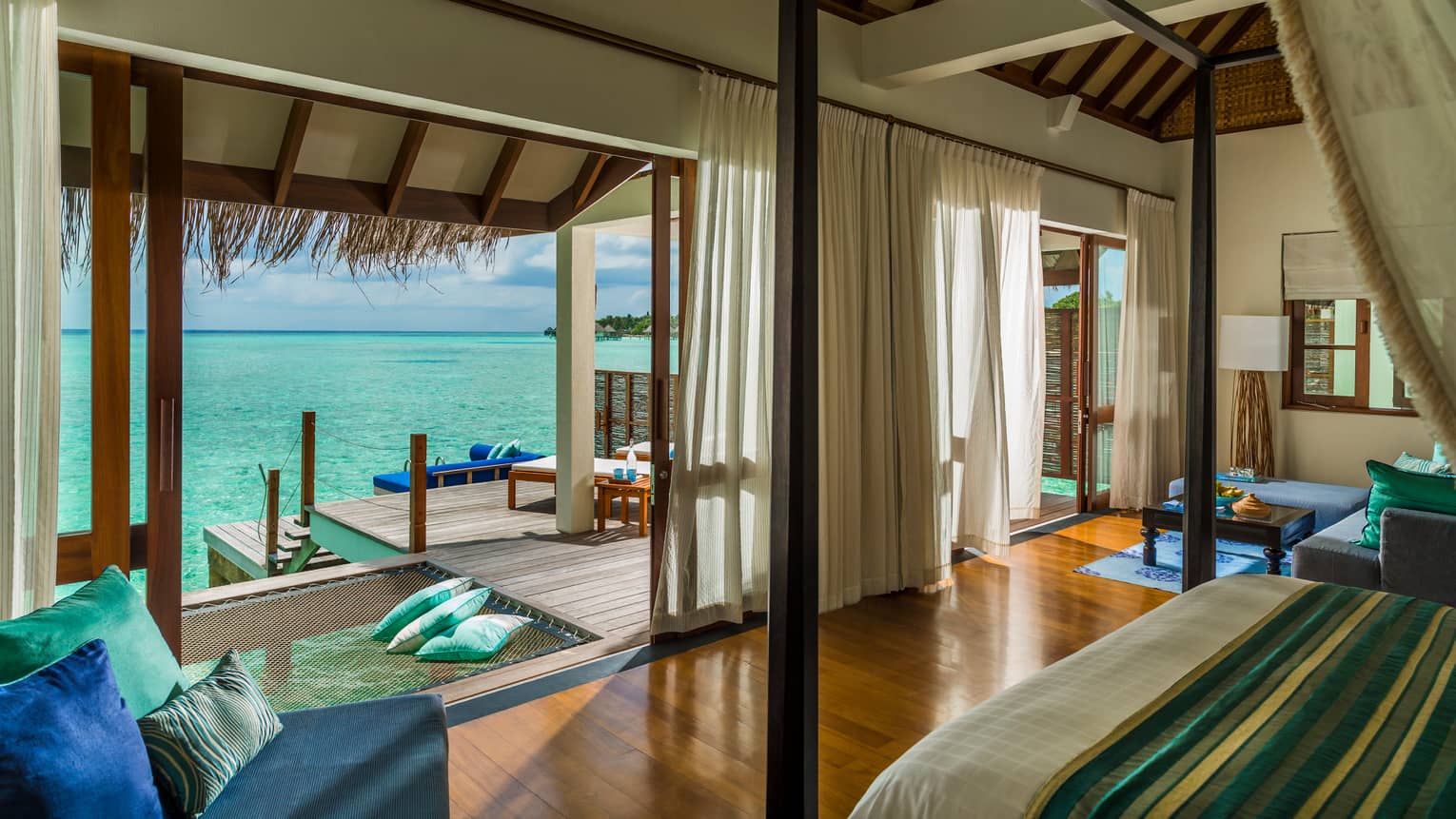 Sunrise Water Villa blue chair with pillows by poster bed across from open wall, patio, ocean views