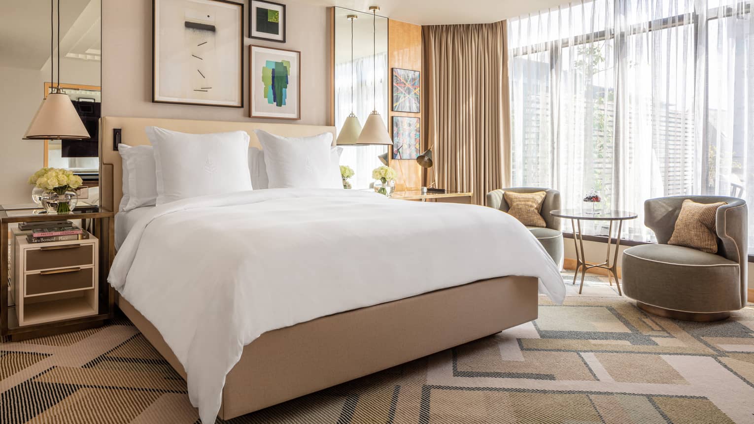 The Junior Conservatory at Four Seasons London Park Lane in Mayfair offers a bright and stylish bedroom space. The room features a large, comfortable bed with crisp white linens centred against a backdrop of modern artwork and a mirrored wall. Two cozy, contemporary armchairs and a small round table sit near floor-to-ceiling windows draped with sheer curtains, providing a relaxed area to enjoy the natural light. The room's decor includes elegant bedside lamps, fresh flowers, and a neutral colour palette with subtle pops of colour from throw pillows, creating a welcoming and sophisticated ambiance.